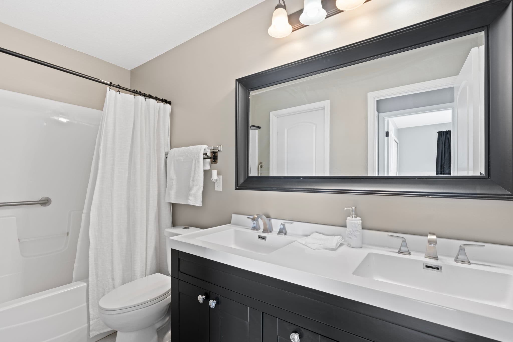 Full bathroom with double-sink vanity, black-framed mirror, and tub-shower with grab bar